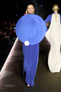 Model walking a runway in a bold blue gown with a large circular blue disc accessory in front, audience visible on sides.