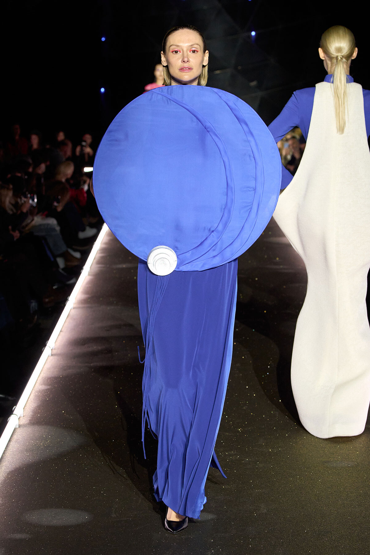 Model walking a runway in a bold blue gown with a large circular blue disc accessory in front, audience visible on sides.