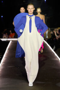 Model walks the runway in a blue long-sleeve top under a loose cream jumpsuit with cape-like panels, white boots, and audience in background.