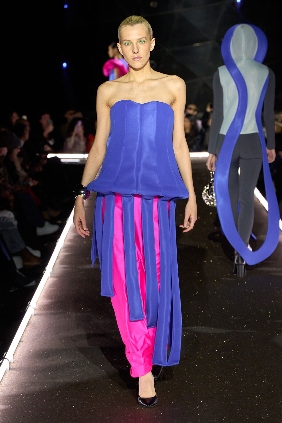 Runway model wearing a cobalt blue strapless top with long blue fringe panels over bright pink pants, walking the catwalk.