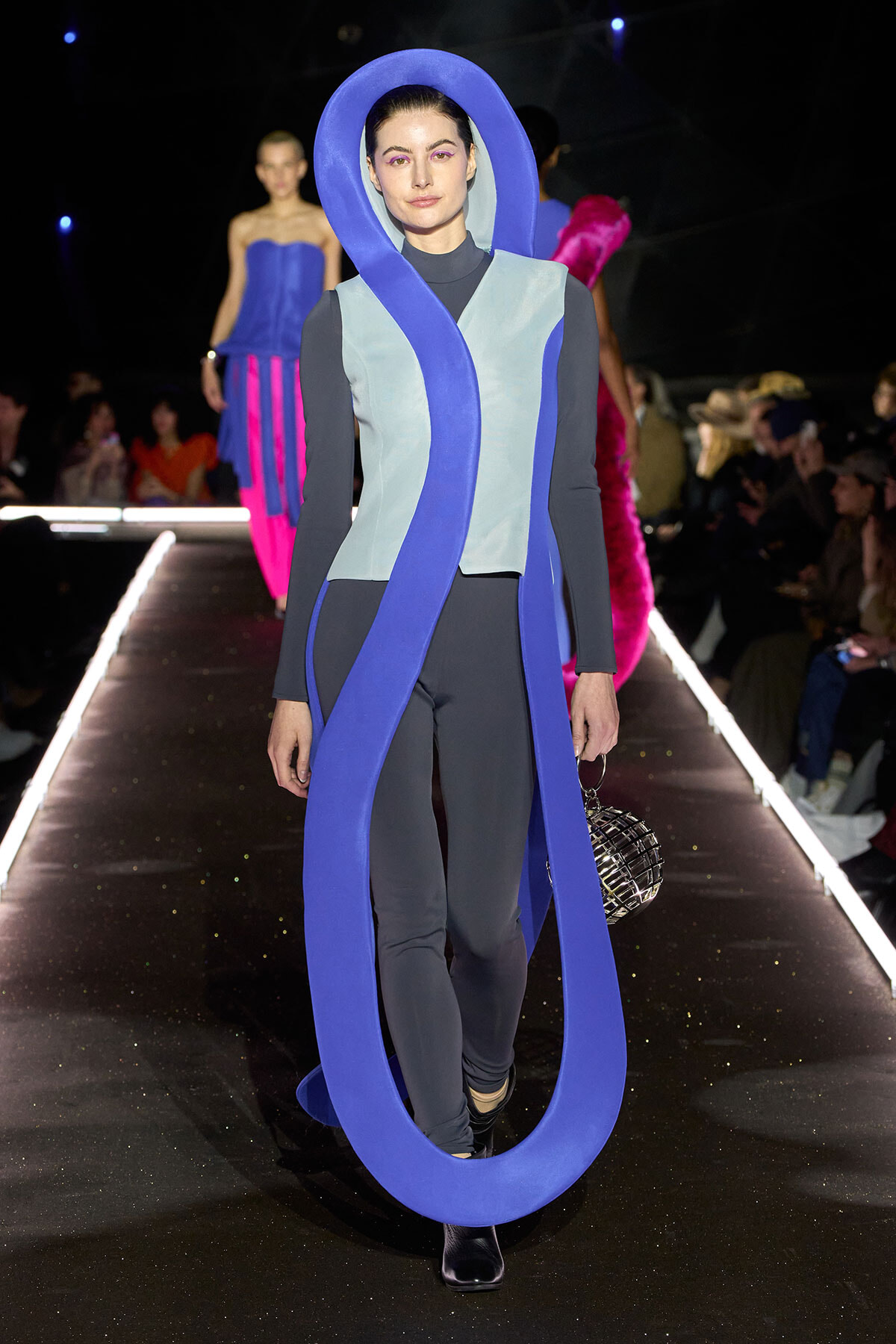 Model walks a fashion runway in a sculptural blue loop costume wrapped around the neck and body, wearing a light blue vest over black sleeves and dark leggings, carrying a metallic spherical handbag.