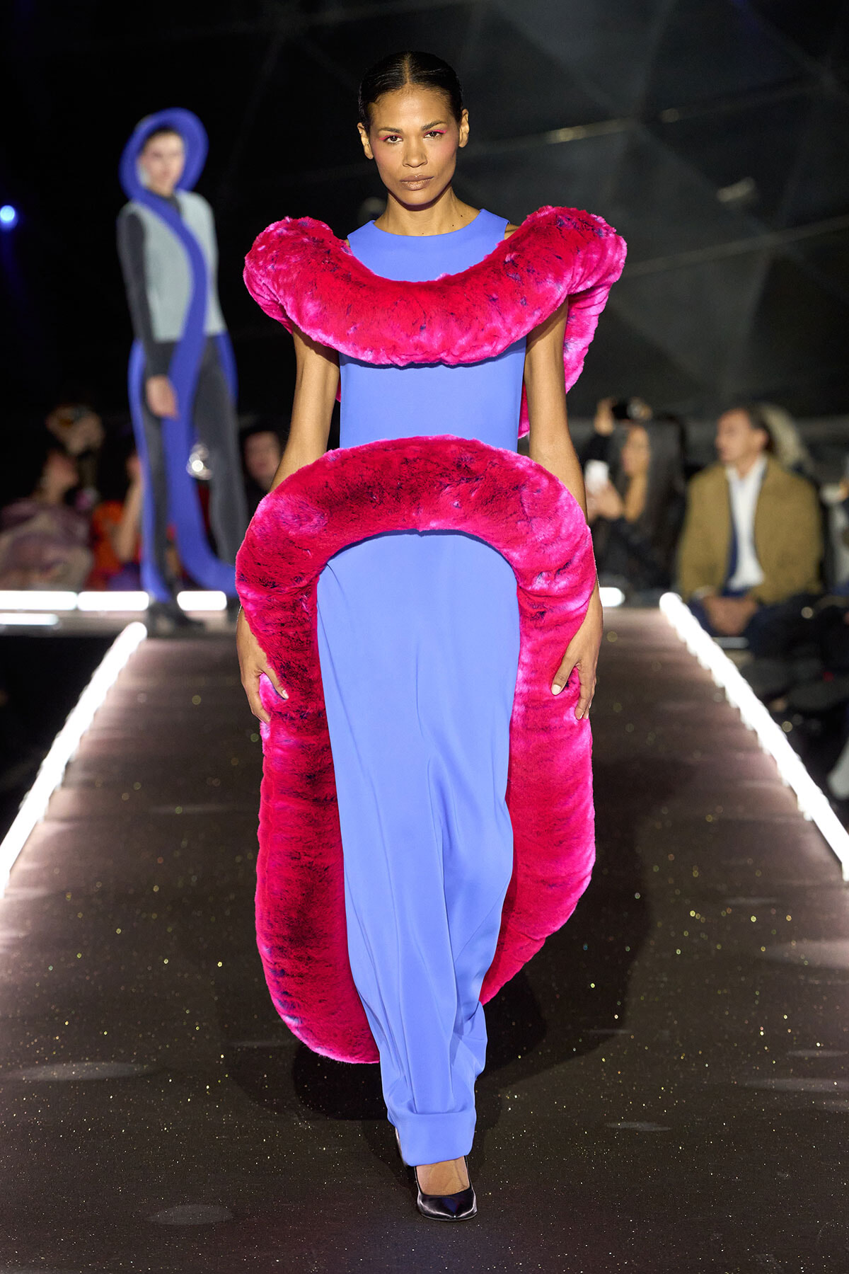 Female model walks a fashion runway in a cobalt-blue dress with oversized pink fur accents around the neck and waist, dramatic makeup visible.