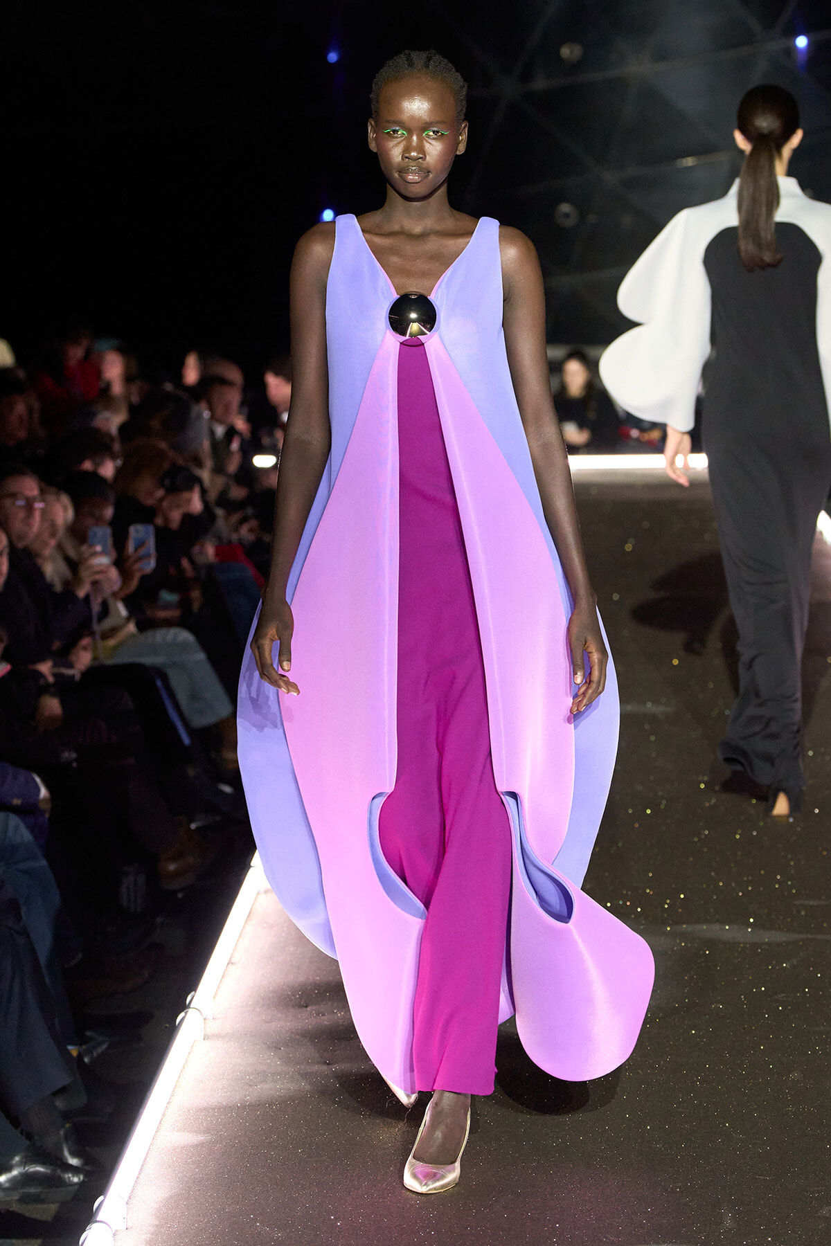 Runway model wearing a flowing lavender and pink color-block dress with a large circular metallic brooch at the chest, audience in background