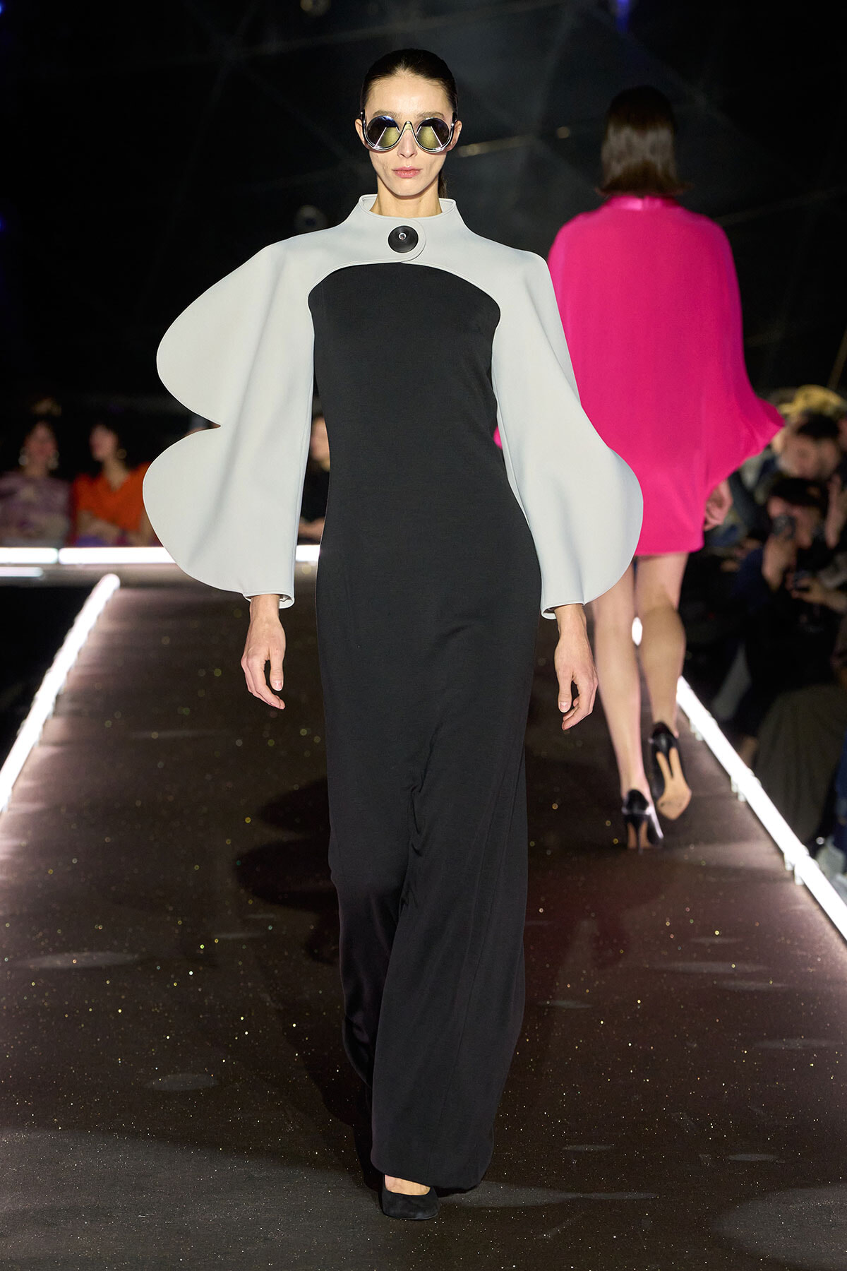 Model walks a runway in a black column dress with oversized white cape sleeves and round sunglasses.”] ,