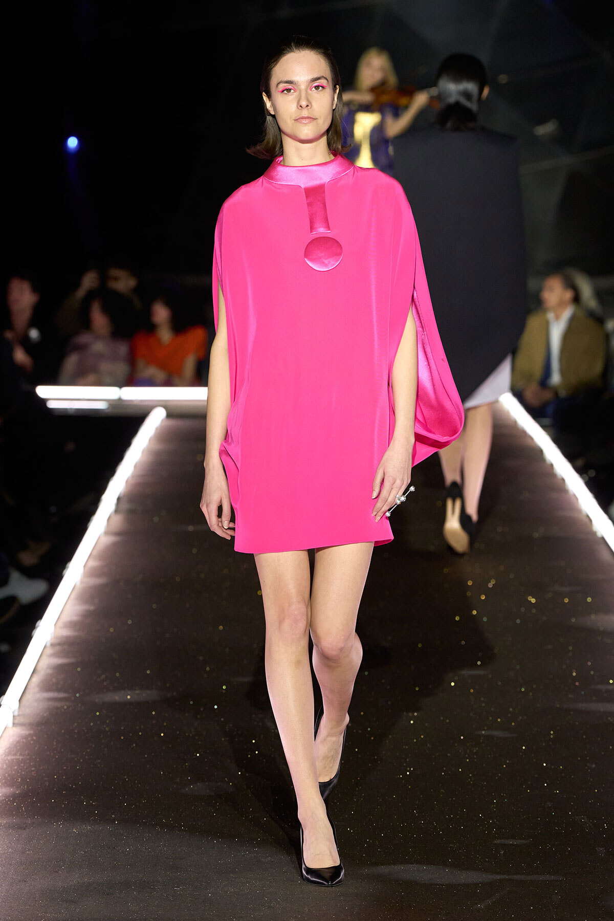 Female model in a hot pink cape-style mini dress with a circular chest detail walks the runway. Audience blurred in the background.