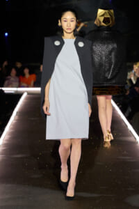 Model walks the runway in a pale gray shift dress layered under a black cape with two large silver buttons at the shoulders side-by-side.