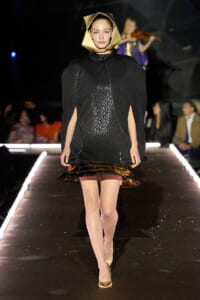 Model walking a runway in a black textured cape with a geometric gold headpiece and metallic gold hem detail. She wears nude heels and a violinist backdrop.