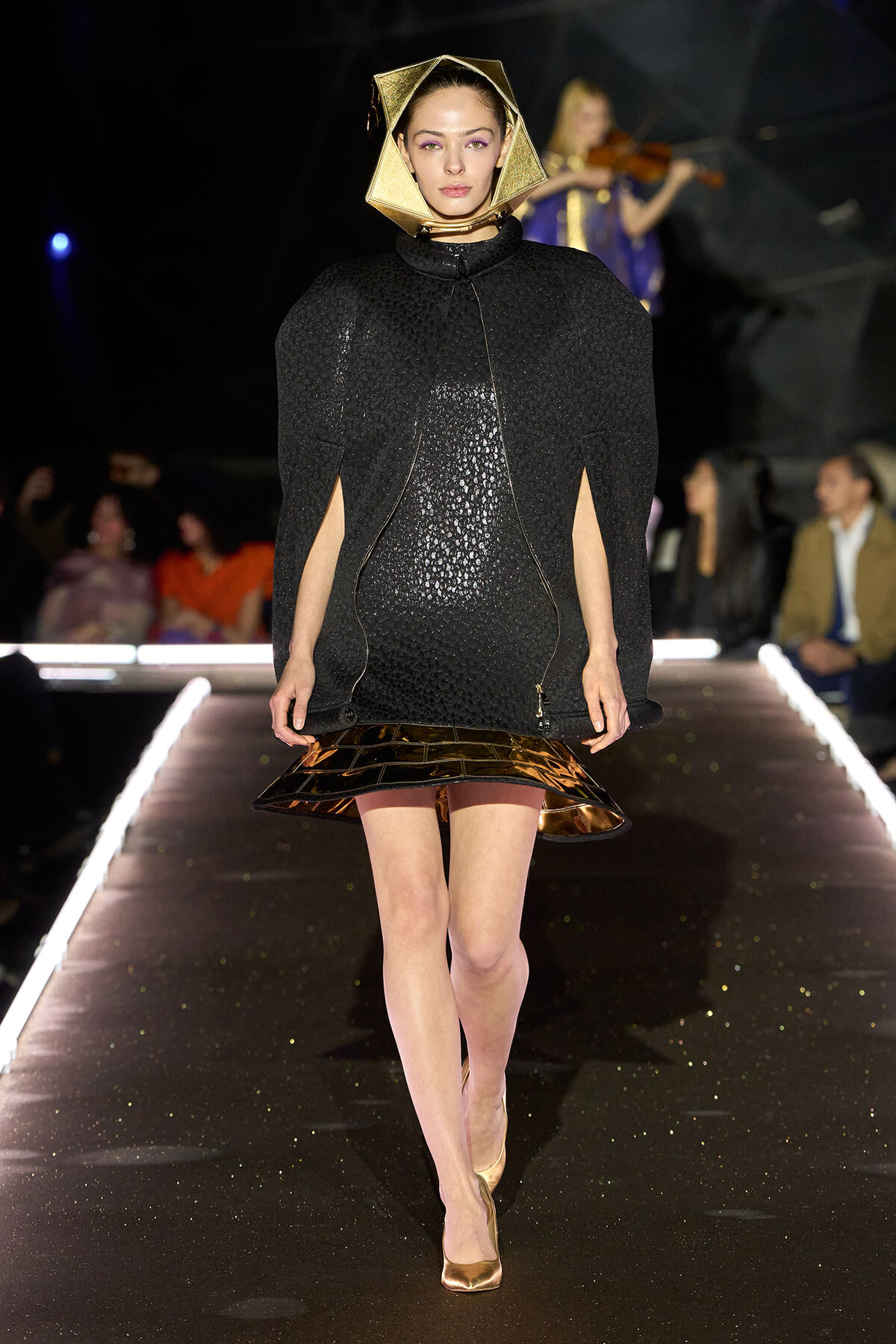 Model walking a runway in a black textured cape with a geometric gold headpiece and metallic gold hem detail. She wears nude heels and a violinist backdrop.