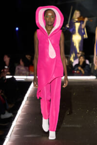 Model walks the runway in a hot-pink sleeveless dress with a large pink hooded collar and matching pants, white ankle boots, and a dark backdrop.