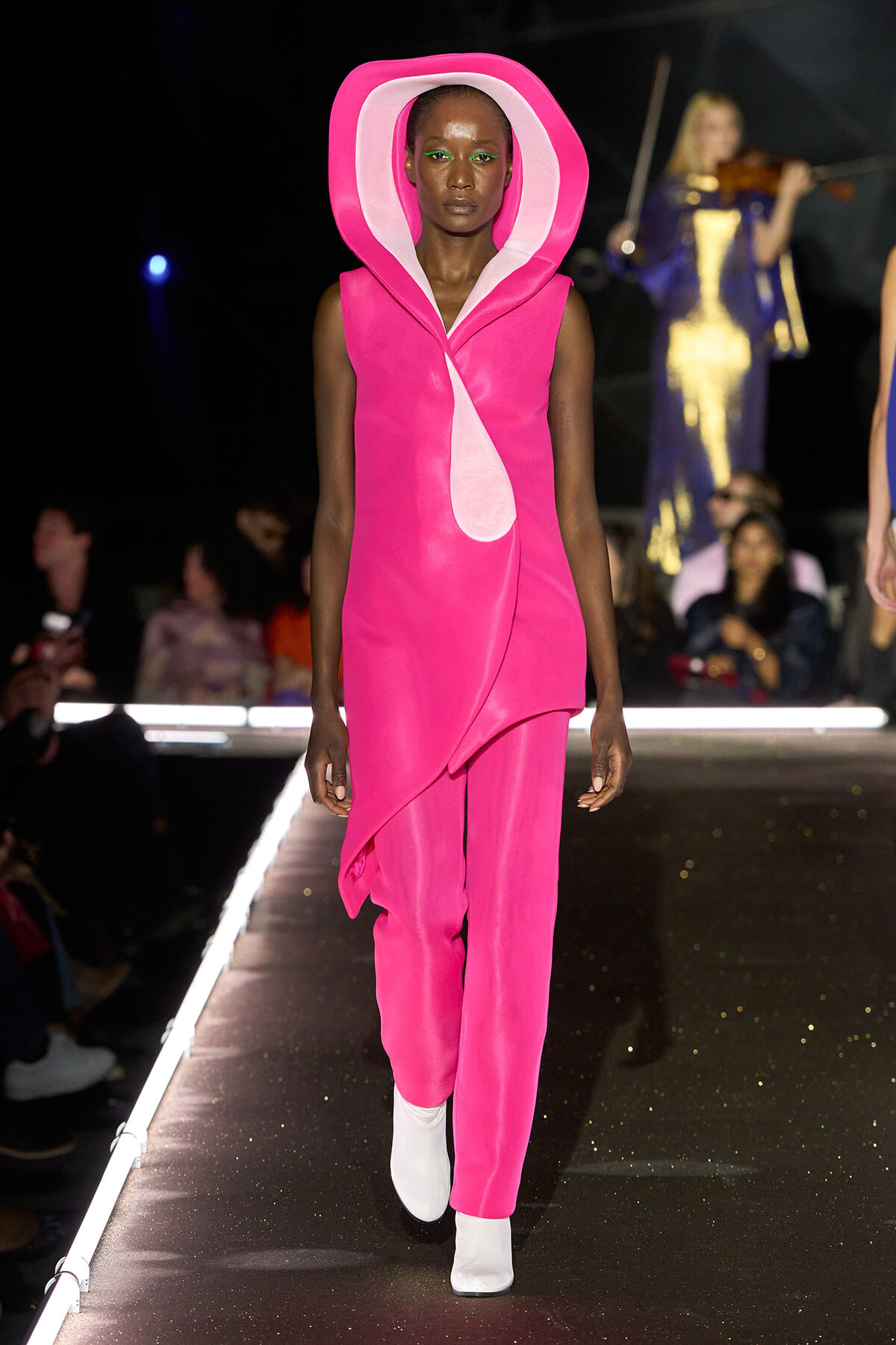 Model walks the runway in a hot-pink sleeveless dress with a large pink hooded collar and matching pants, white ankle boots, and a dark backdrop.