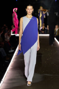 Model walking a fashion show wearing a sleeveless, color-blocked asymmetric blue and light gray top over wide pale trousers on a glossy runway. Audience in dim backdrop.