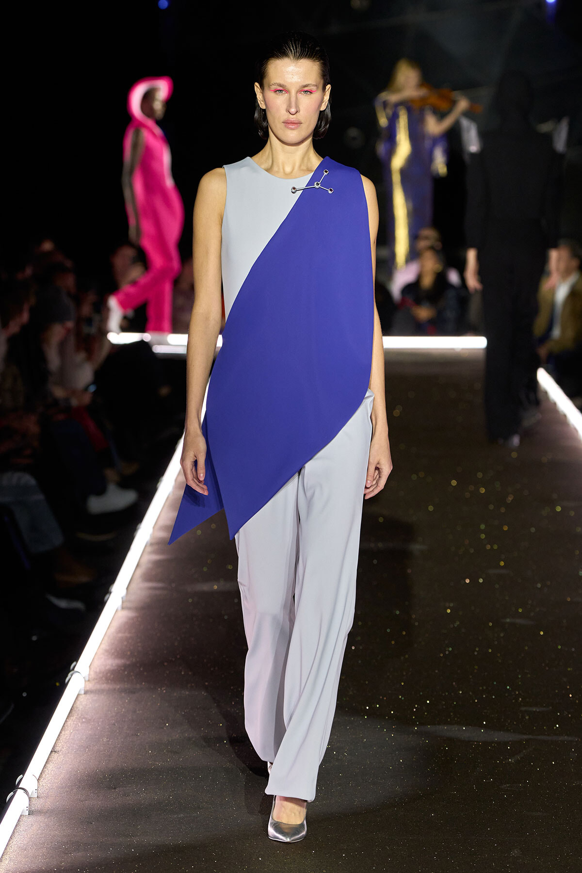 Model walking a fashion show wearing a sleeveless, color-blocked asymmetric blue and light gray top over wide pale trousers on a glossy runway. Audience in dim backdrop.