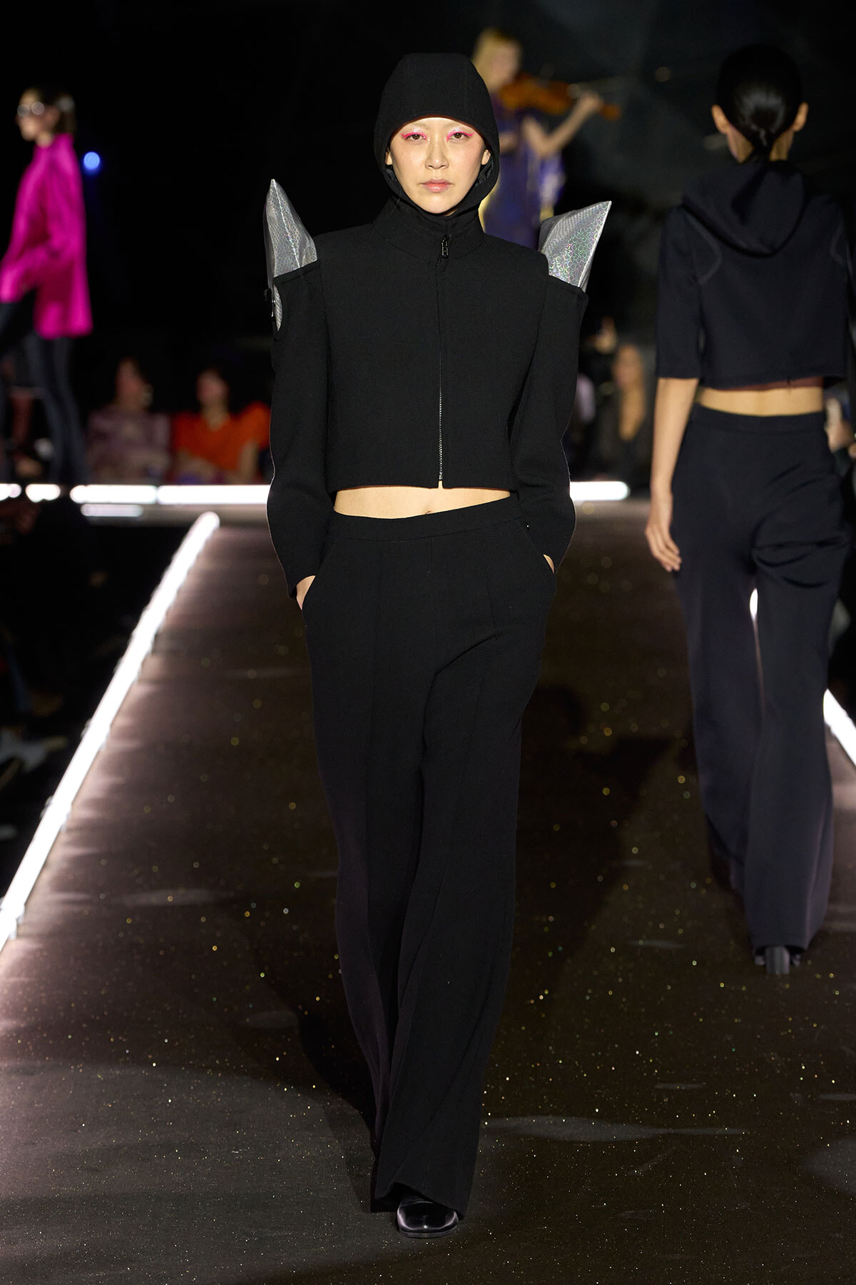 Model walking a fashion runway in an all-black hooded outfit with a cropped zip-front top and wide-leg pants, featuring angular silver shoulder accents.