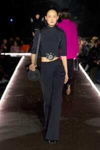 Model on a runway wearing a navy cropped top and wide-leg pants, with a circular beaded belt and round black purse.
