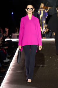 Model on a fashion runway wearing a bright pink oversized coat, black pants, black heels, large sunglasses, and a small black handbag.