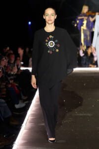 Model walking a fashion show in a black ensemble with a colorful circular gem design on the chest edge of the sweater/pullover