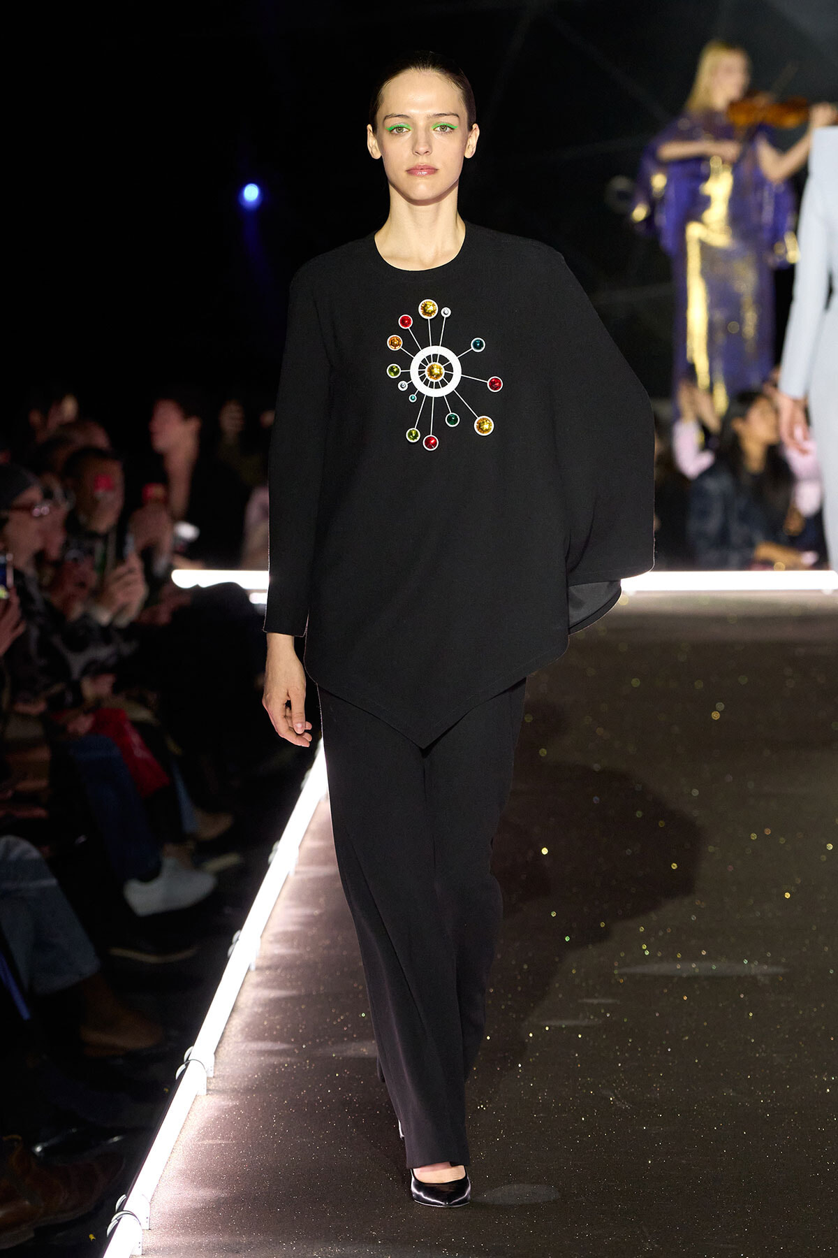 Model walking a fashion show in a black ensemble with a colorful circular gem design on the chest edge of the sweater/pullover
