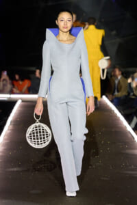 Model walking the runway in a pale blue, structured jumpsuit with blue interior, holding a white circular cage handbag.