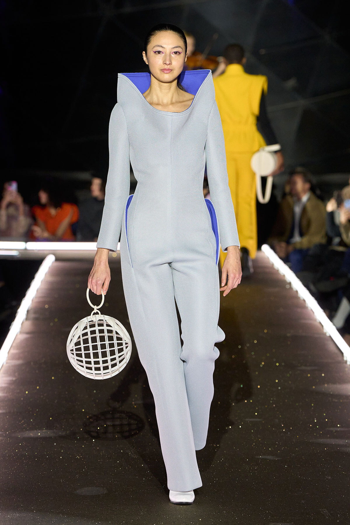 Model walking the runway in a pale blue, structured jumpsuit with blue interior, holding a white circular cage handbag.