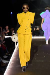 Runway model in a bright yellow sculptural suit with oversized shoulders, black sleeves, belt, and a circular white handbag.