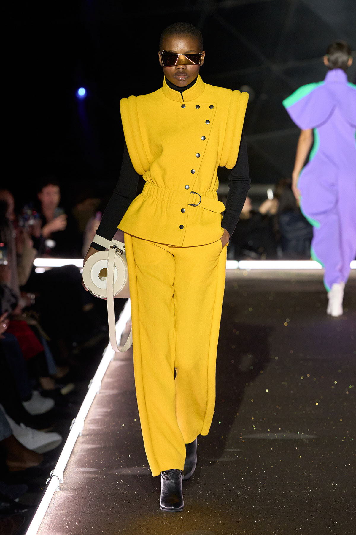 Runway model in a bright yellow sculptural suit with oversized shoulders, black sleeves, belt, and a circular white handbag.