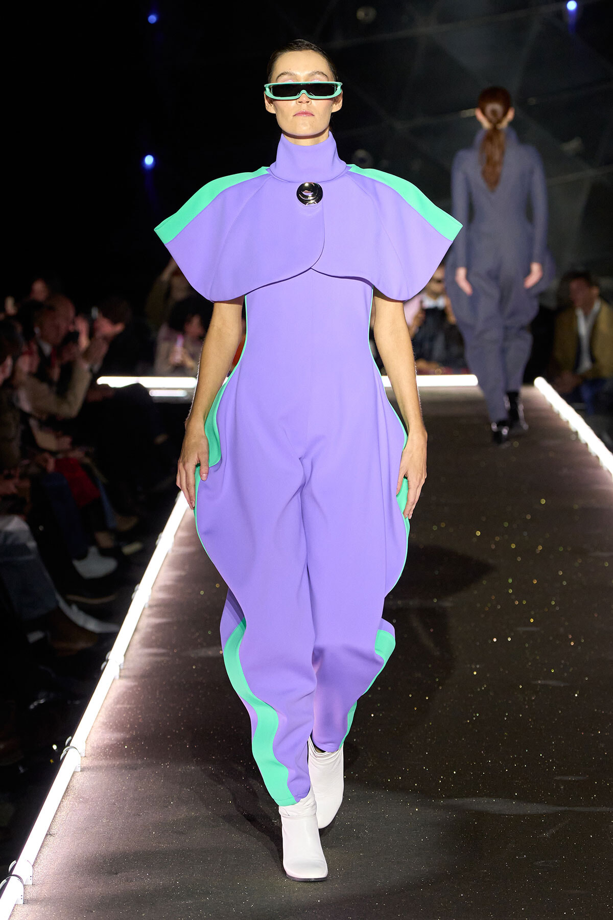 Model walks a fashion runway wearing a lavender jumpsuit with mint green panels and oversized cape-like shoulders, white boots, and futuristic sunglasses.