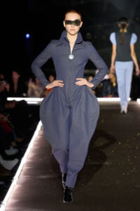 Model walks the runway in a navy blue zip-up jumpsuit with oversized hip pockets, paired with futuristic sunglasses and black ankle boots.