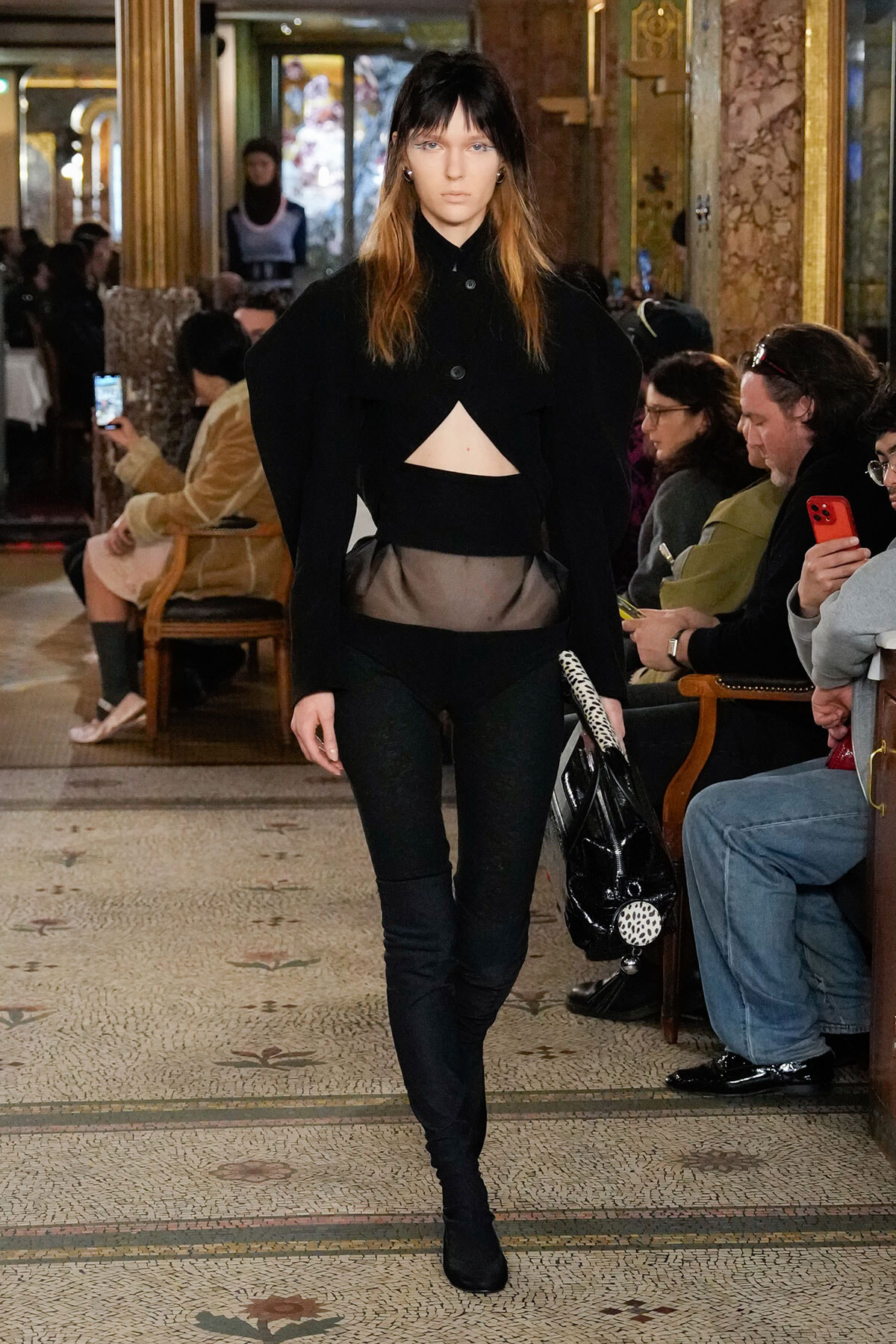 Runway model wearing an all-black outfit with a cropped midriff triangle, walking down a fashion show aisle in an ornate venue with spectators.