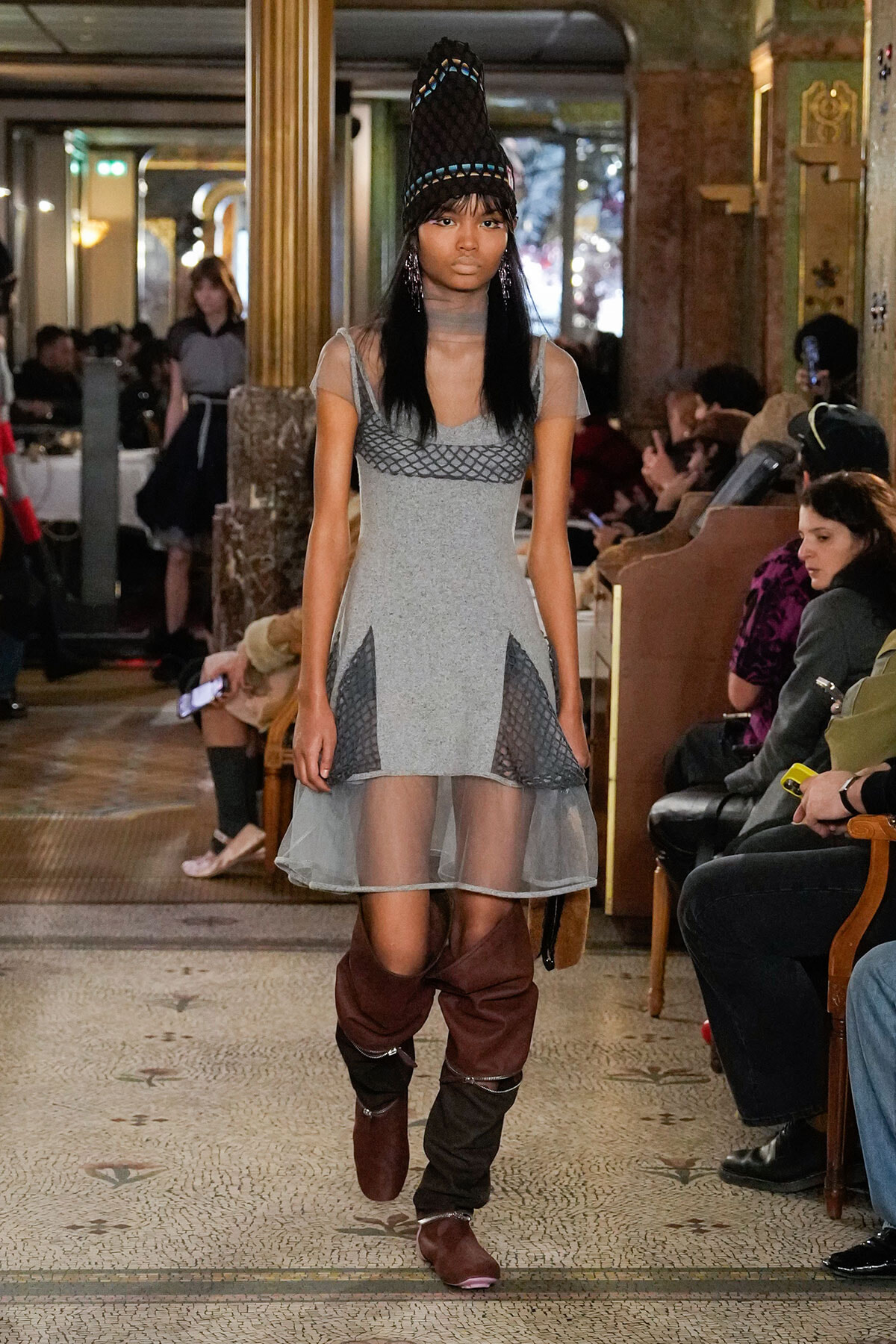 Model walking a runway in a gray mini dress with mesh panels and a sheer hem, paired with a tall sculptural black hat with blue accents and brown knee-high boots.