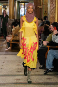 Model walks a runway in a bright yellow dress with red graphics, paired with a pink headwrap and boots in an ornate venue.