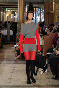 Model walks a runway in a gray and red color-block outfit with a tall black cone hat and black boots, carrying a black bag.