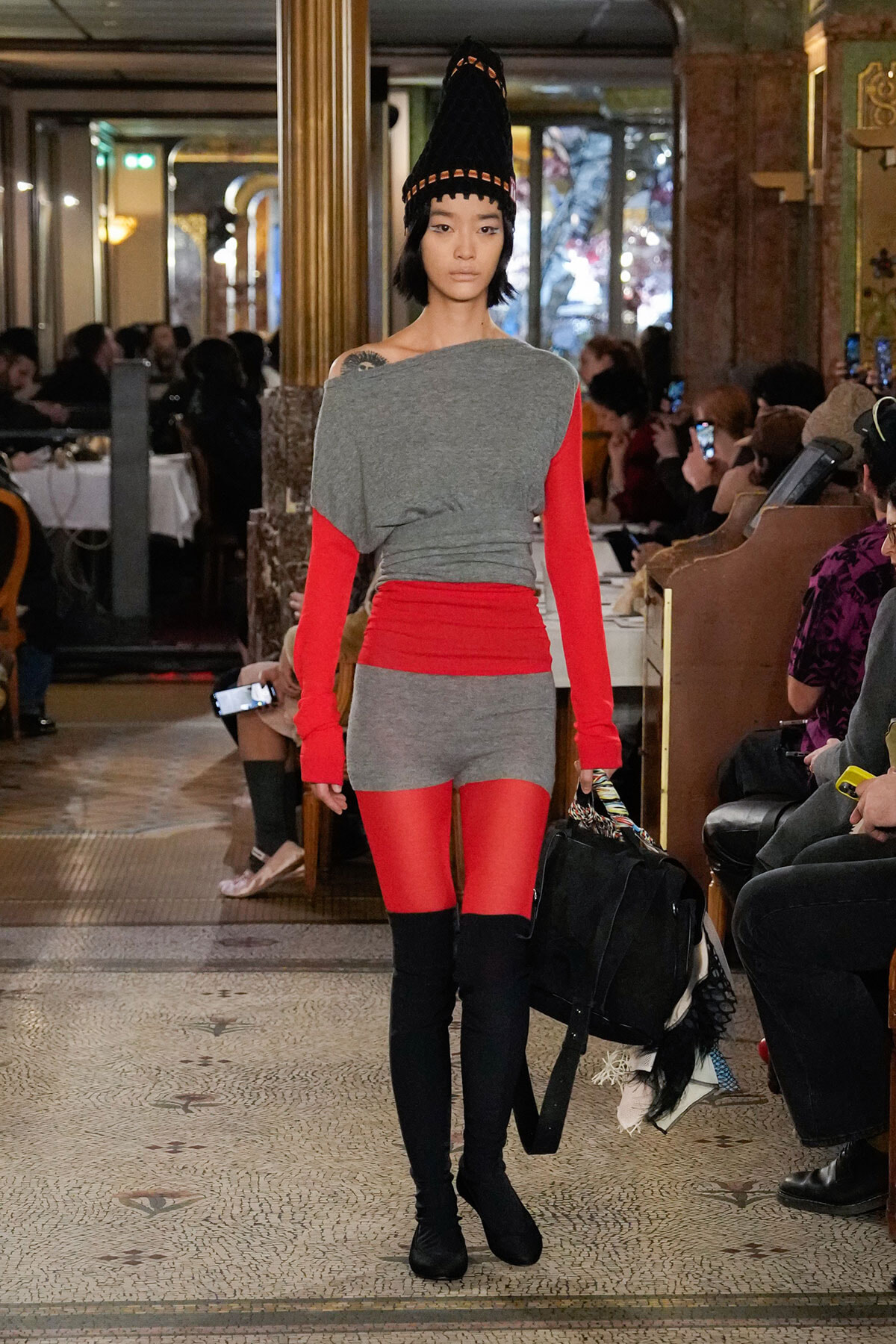 Model walks a runway in a gray and red color-block outfit with a tall black cone hat and black boots, carrying a black bag.