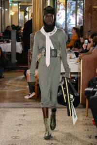 Model walks the runway in a sage-green jumpsuit with an oversized belt, white scarf, and green ankle boots, carrying a large patterned tote and fabrics.