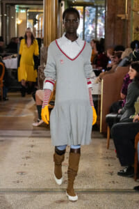 Model walking a runway in a light gray sweater dress with red V-neck trim, white striped shirt underneath, yellow gloves, and slouchy brown boots; audience in a stylish venue backdrop.