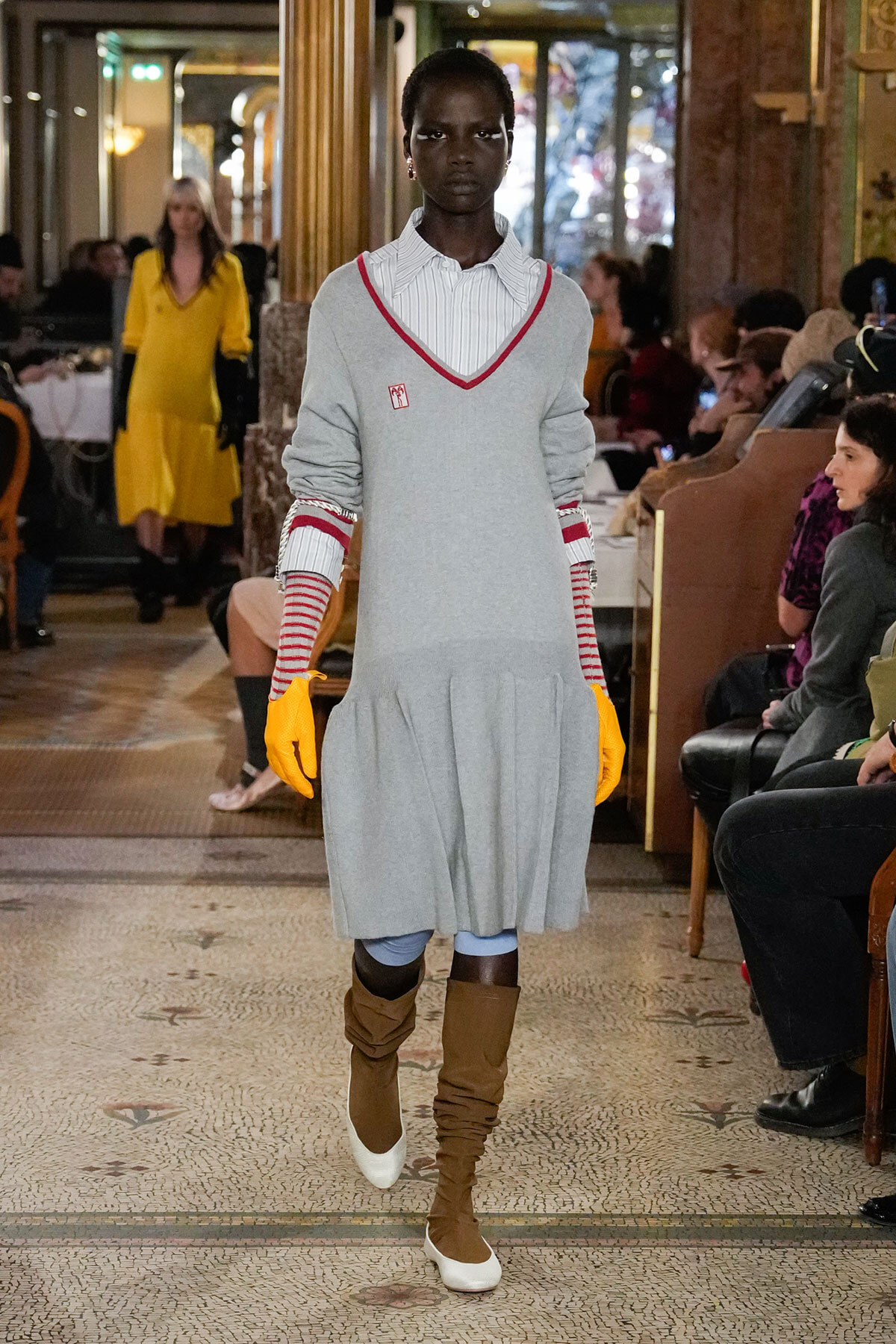 Model walking a runway in a light gray sweater dress with red V-neck trim, white striped shirt underneath, yellow gloves, and slouchy brown boots; audience in a stylish venue backdrop.