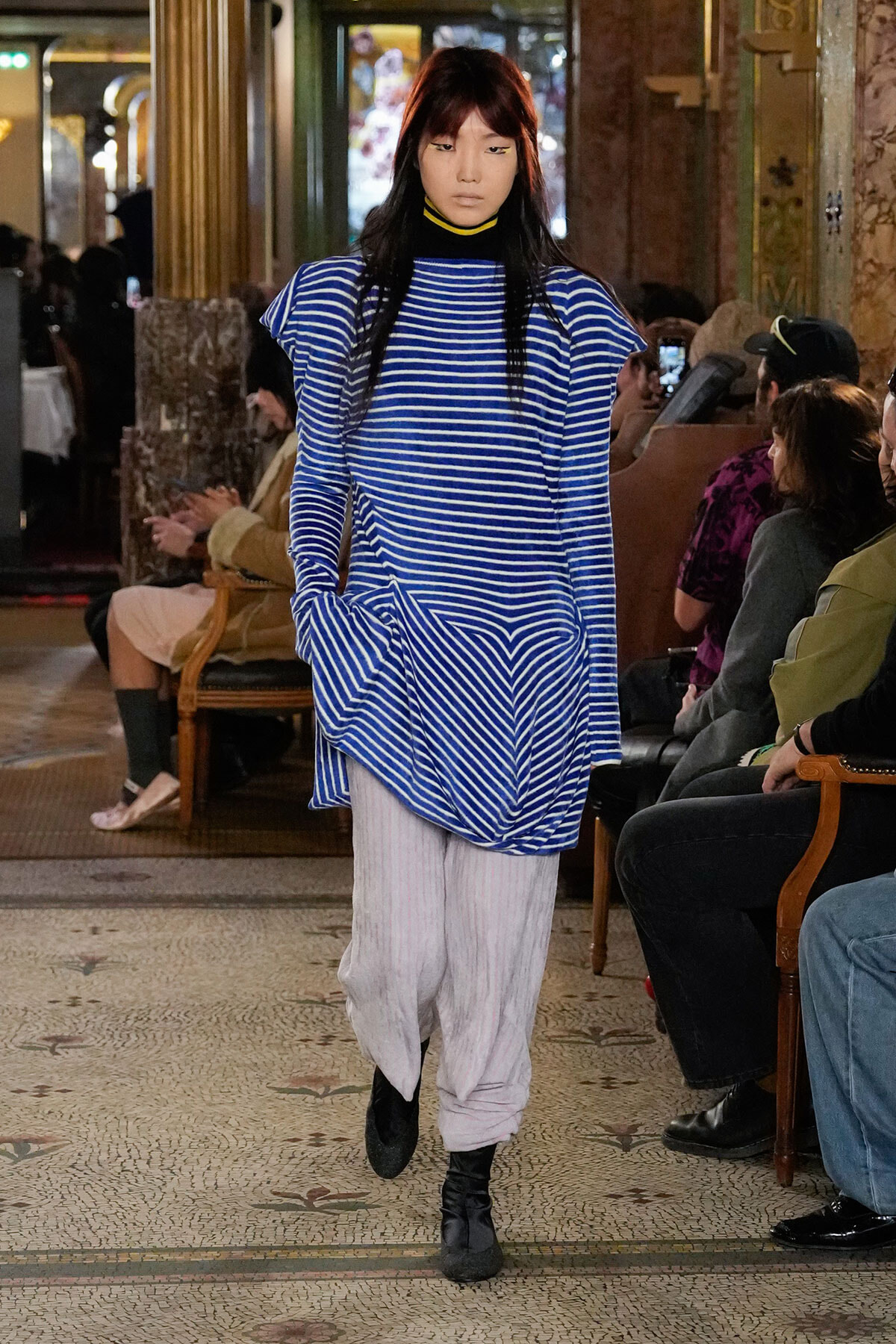 Model walks the runway in a blue and white striped oversized top with large front pockets, paired with light gray loose pants and black ankle boots, audience seated in ornate venue behind.