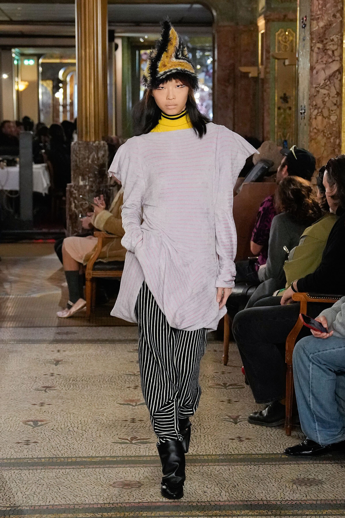 Model walks a runway wearing an oversized pale pink top, black-and-white vertical striped pants, and knee-high black boots with a yellow-and-black feathered headpiece sanely perched on her head in an ornate venue.