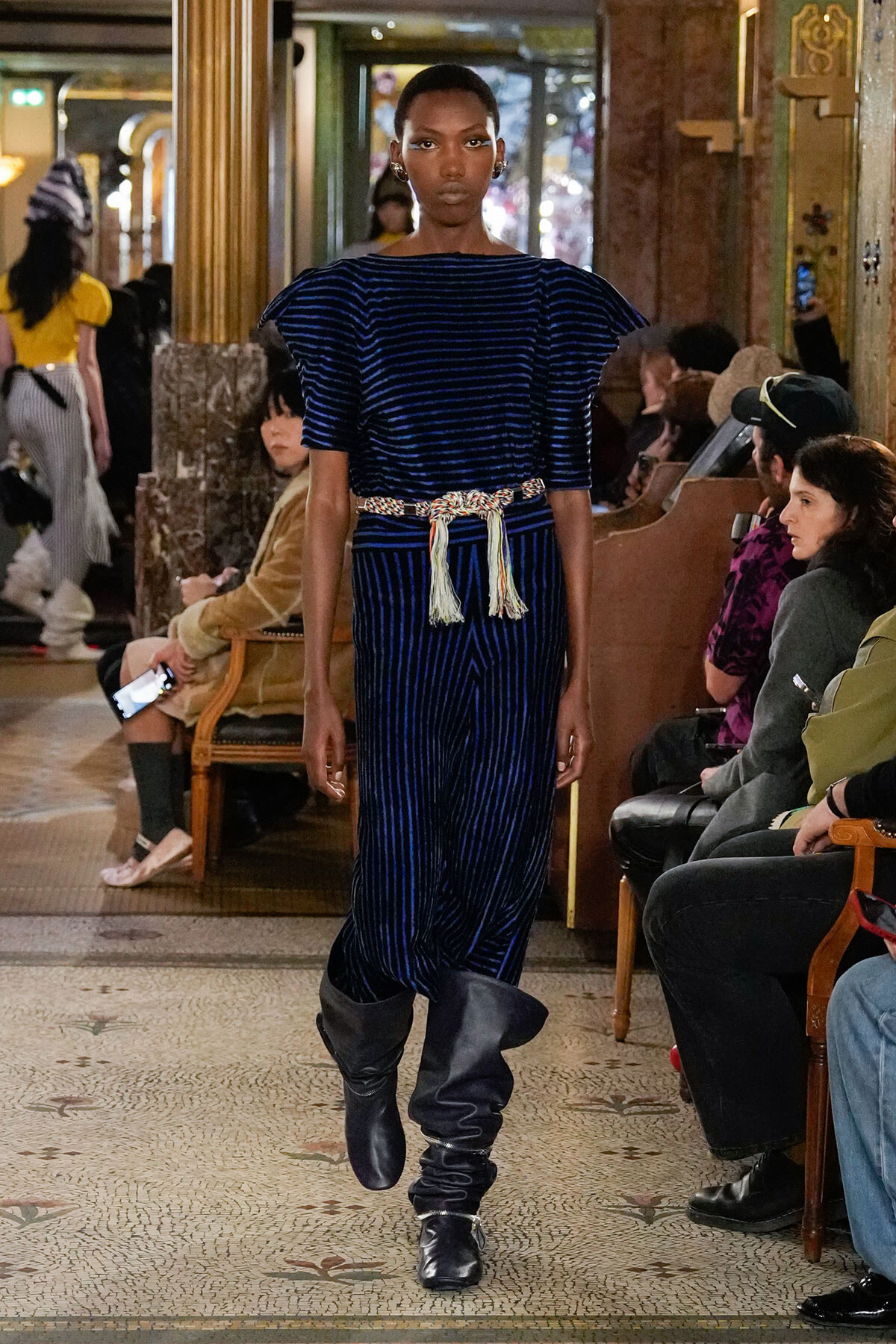 Model walks a fashion show wearing a navy blue velvet striped outfit with a braided belt and tall slouch boots inside an ornate venue.