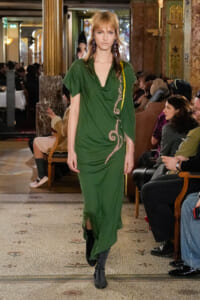 Model wearing a green draped dress with pink embroidery walks down a runway in an ornate venue with seated audience.