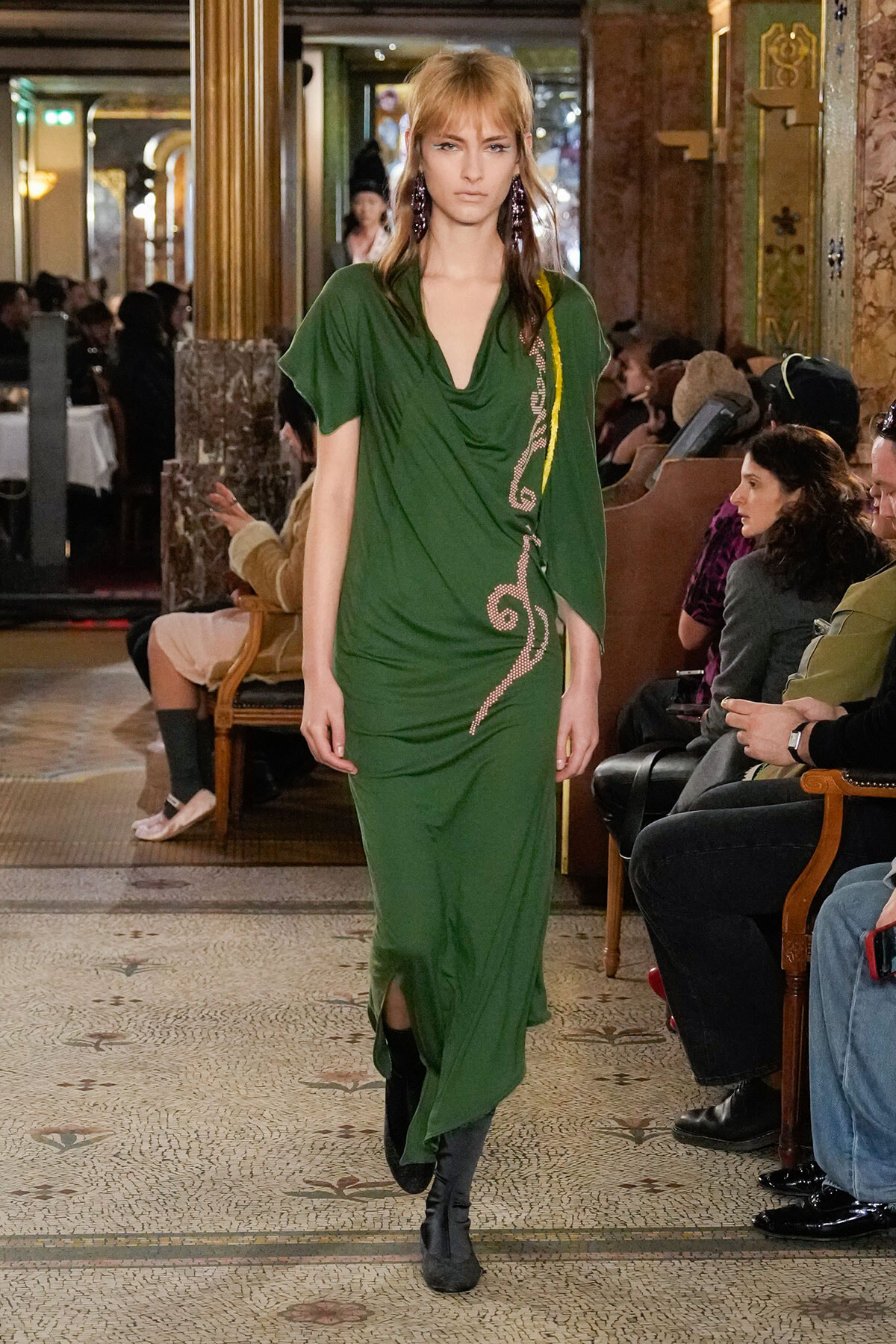 Model wearing a green draped dress with pink embroidery walks down a runway in an ornate venue with seated audience.