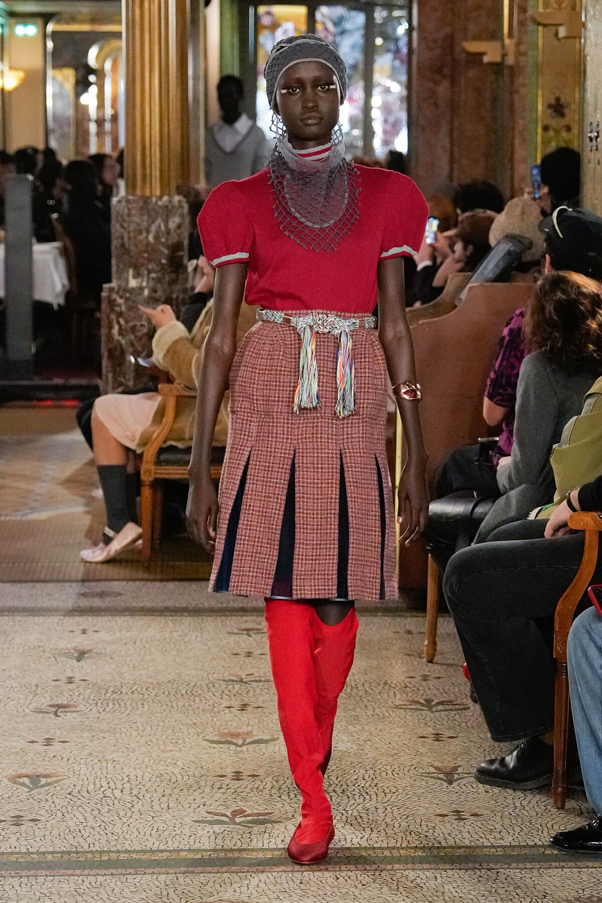 Model walks a runway in a red top, plaid pleated skirt with vertical panels, red knee-high boots, and layered necklace with a headwrap; audience seated along the sides.