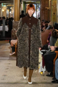 Model walks a fashion show wearing a long textured gray fur coat with brown panels, brown boots, and white shoes, carrying a light bag in a grand, ornate venue with seated audience.