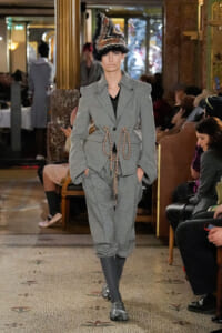 Model walks a fashion show in a gray textured suit with an braided rope belt and a dramatic feathered hat in an ornate venue.