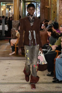 Female model walks a runway in a brown double-breasted coat with rope ties, distressed gray jeans with paint splatters, and burgundy boots, carrying a netted bag.