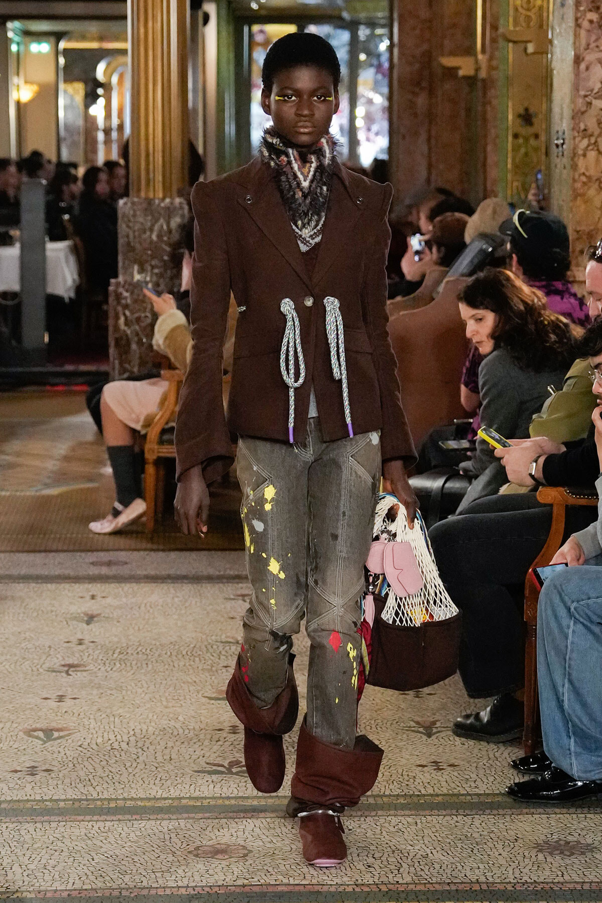 Female model walks a runway in a brown double-breasted coat with rope ties, distressed gray jeans with paint splatters, and burgundy boots, carrying a netted bag.