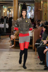 Model walks the runway in a gray denim jacket with paint splatters, red top and tights, and tall black boots, inside an ornate venue.