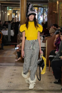 Model walks a runway in a bright yellow top with black trim and high-waisted black-and-white striped pants, topped by a tall furry hat when lighting catches the stage.