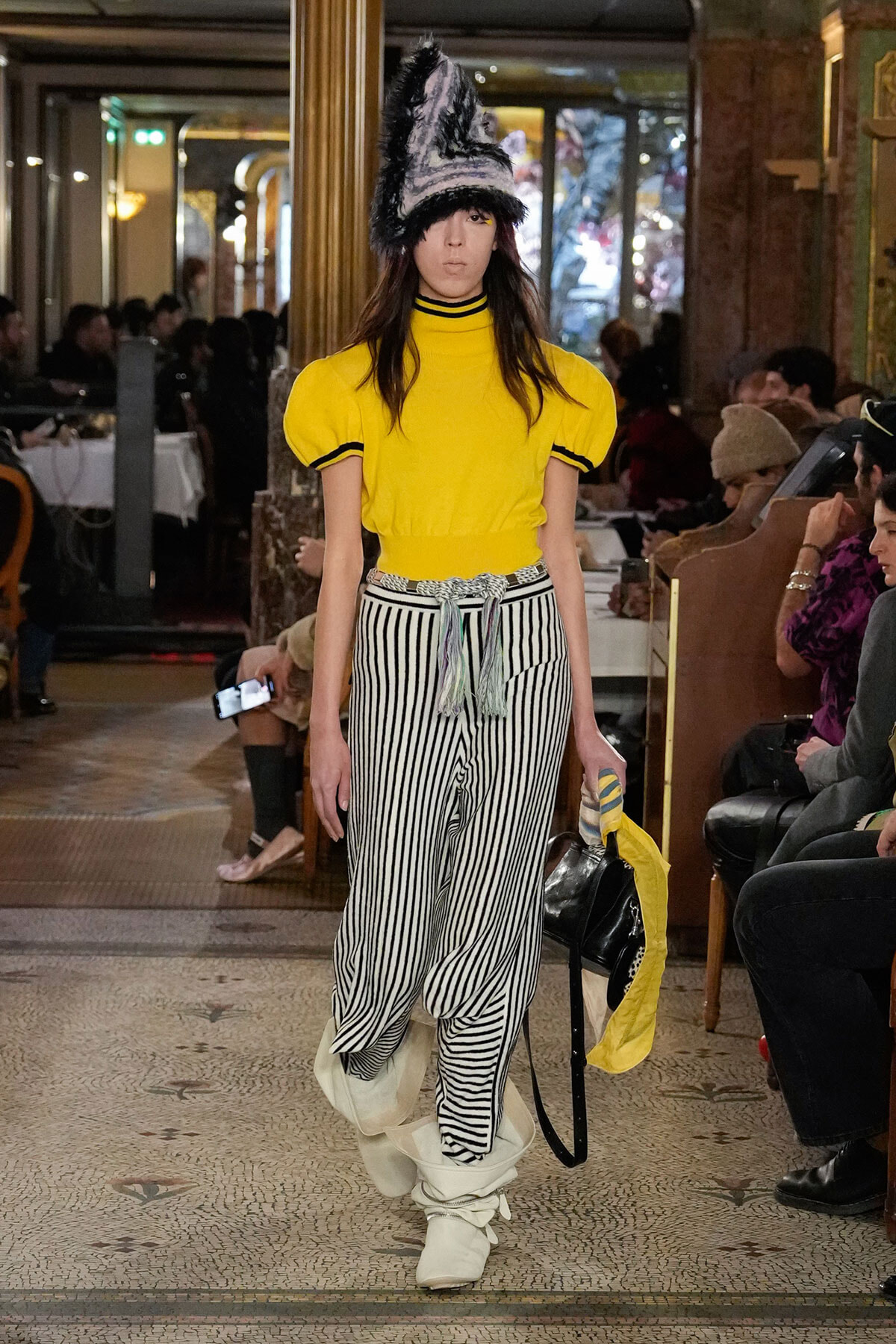 Model walks a runway in a bright yellow top with black trim and high-waisted black-and-white striped pants, topped by a tall furry hat when lighting catches the stage.