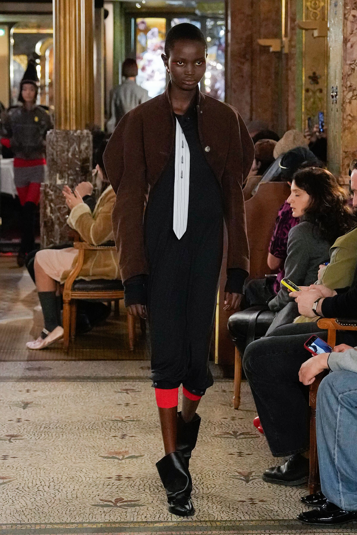 Model on a runway wearing a brown jacket, white striped tie, black pants with red cuffs, and black boots in a luxurious, ornate venue with seated audience.