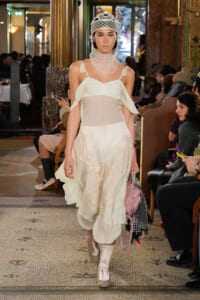 Model walks a runway in an ivory layered dress with shoulder straps and a sculptural mesh neck/hood, indoors at a fashion show.